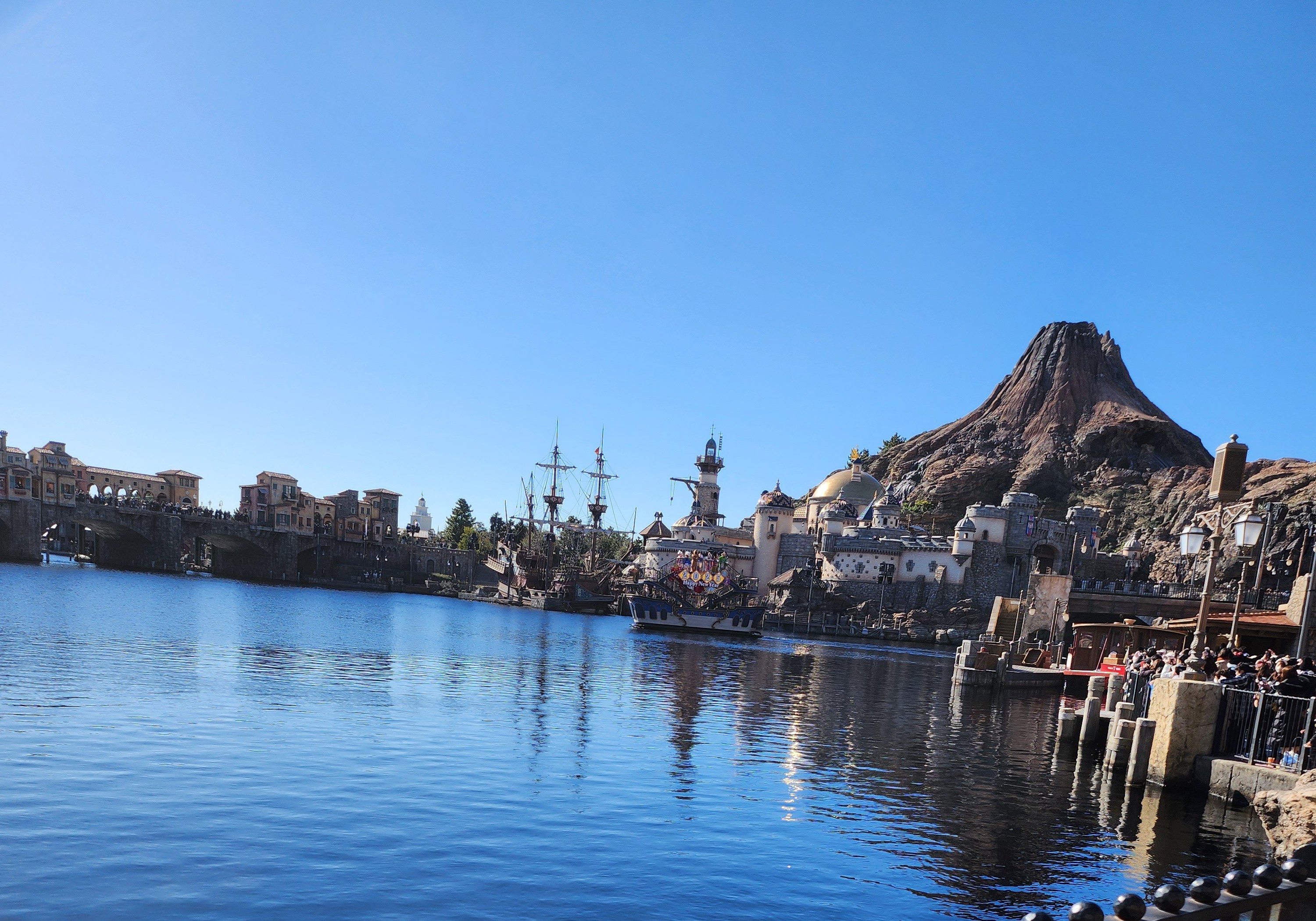 Daytime view across Tokyo DisneySea Mediterranean Harbor, with ships, the harbor town, and Mount Prometheus in the background.