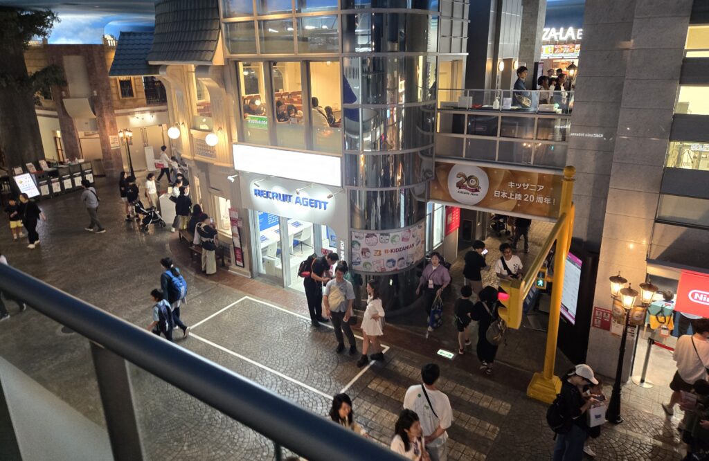 A street inside KidZania Tokyo indoor mini city, with the RECRUIT AGENT pavilion, a glass elevator, and a 20th anniversary banner.