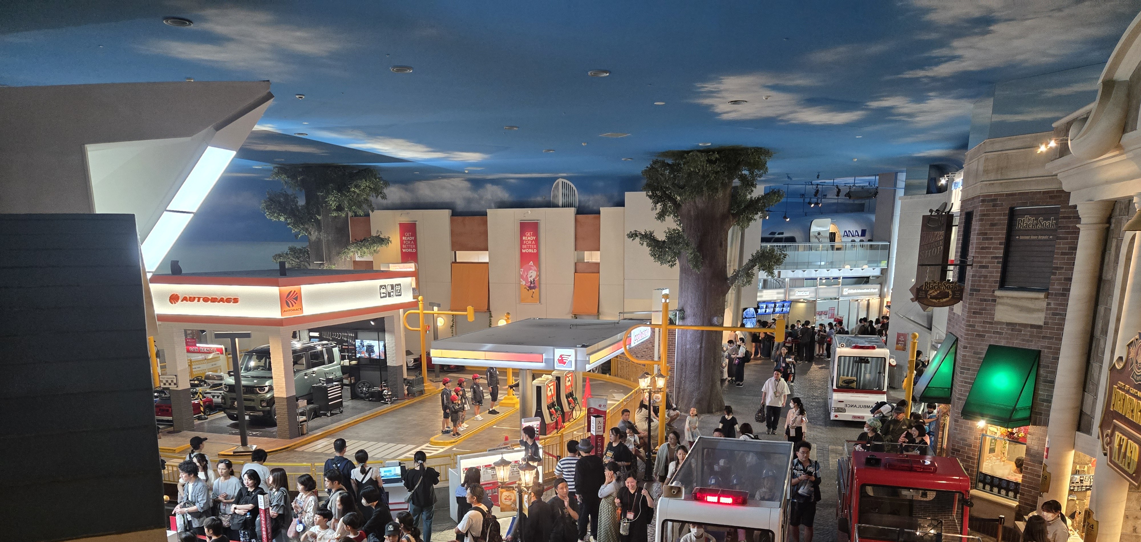 A wide street inside KidZania Tokyo lined with an AUTOBACS car-service pavilion, a fuel pump, fire trucks, and an ambulance-style vehicle.