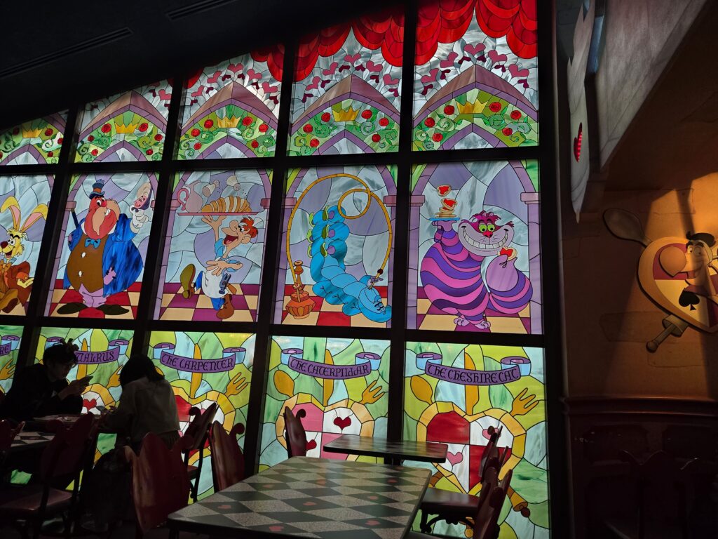 Alice in Wonderland themed stained glass windows inside Queen of Hearts Banquet Hall at Tokyo Disneyland, with mostly empty tables.