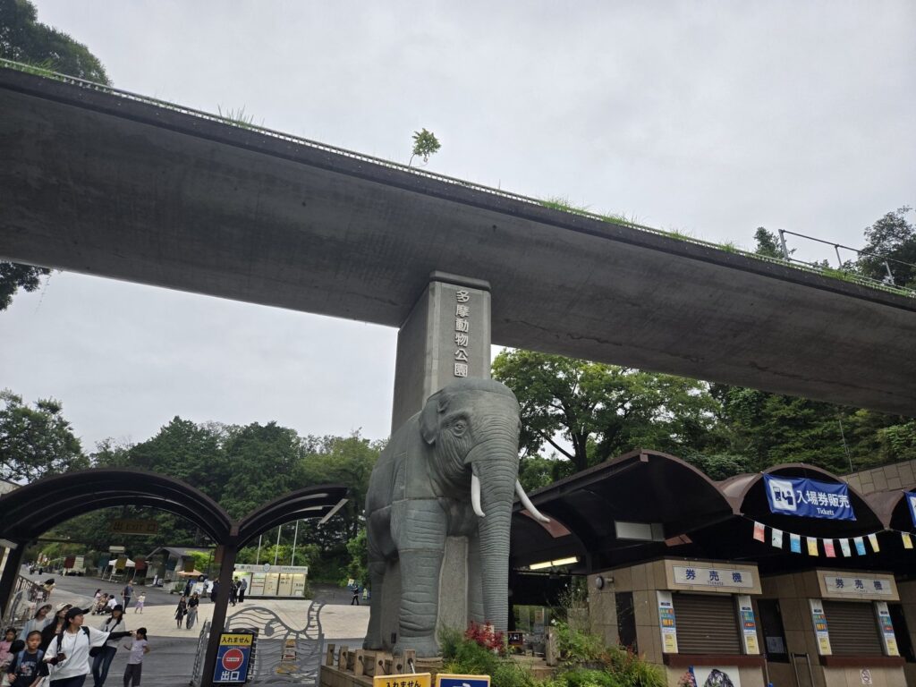 Stone elephant monument marking the entrance to Tama Zoological Park, with the Tama Monorail passing overhead.
