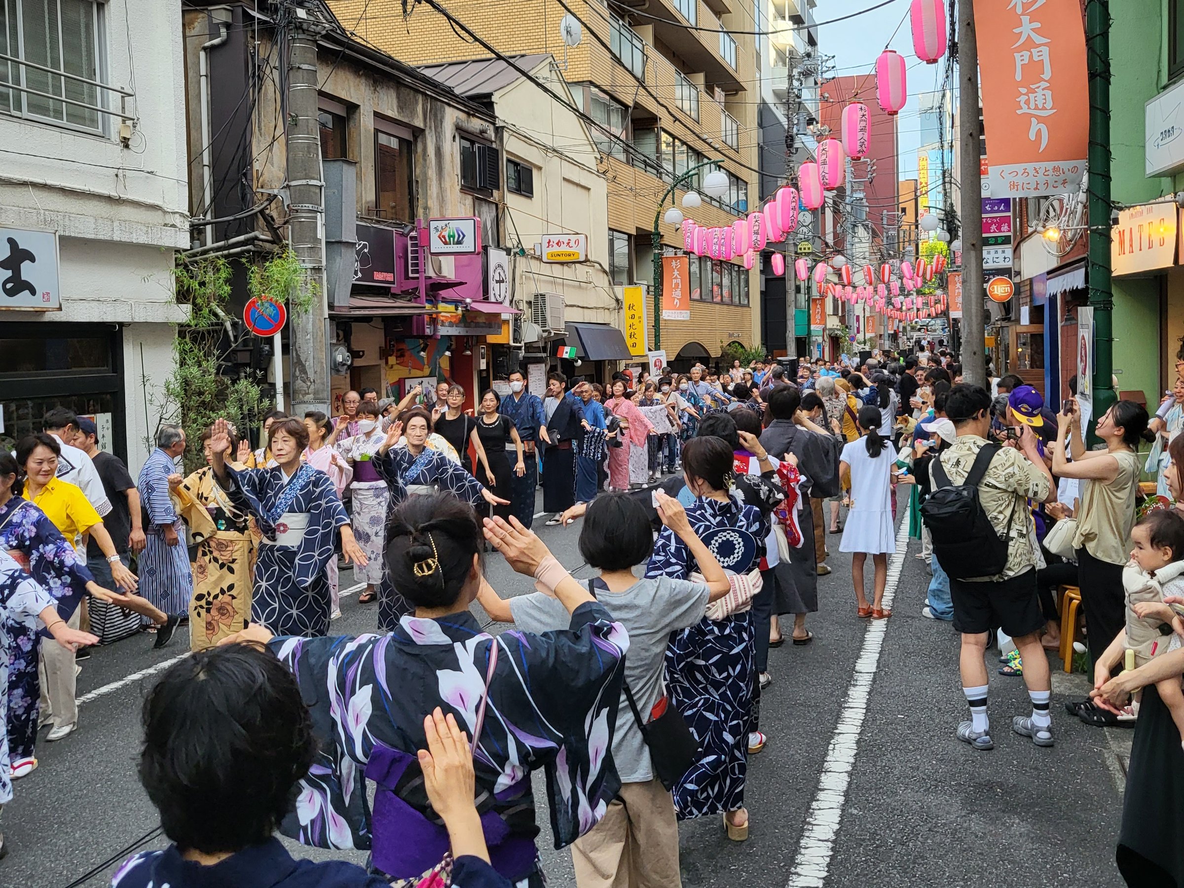 Dancers in yukata moving in a line down a narrow street lined with pink paper lanterns during the Sugi-Daimon-Dori bon odori in Tokyo.