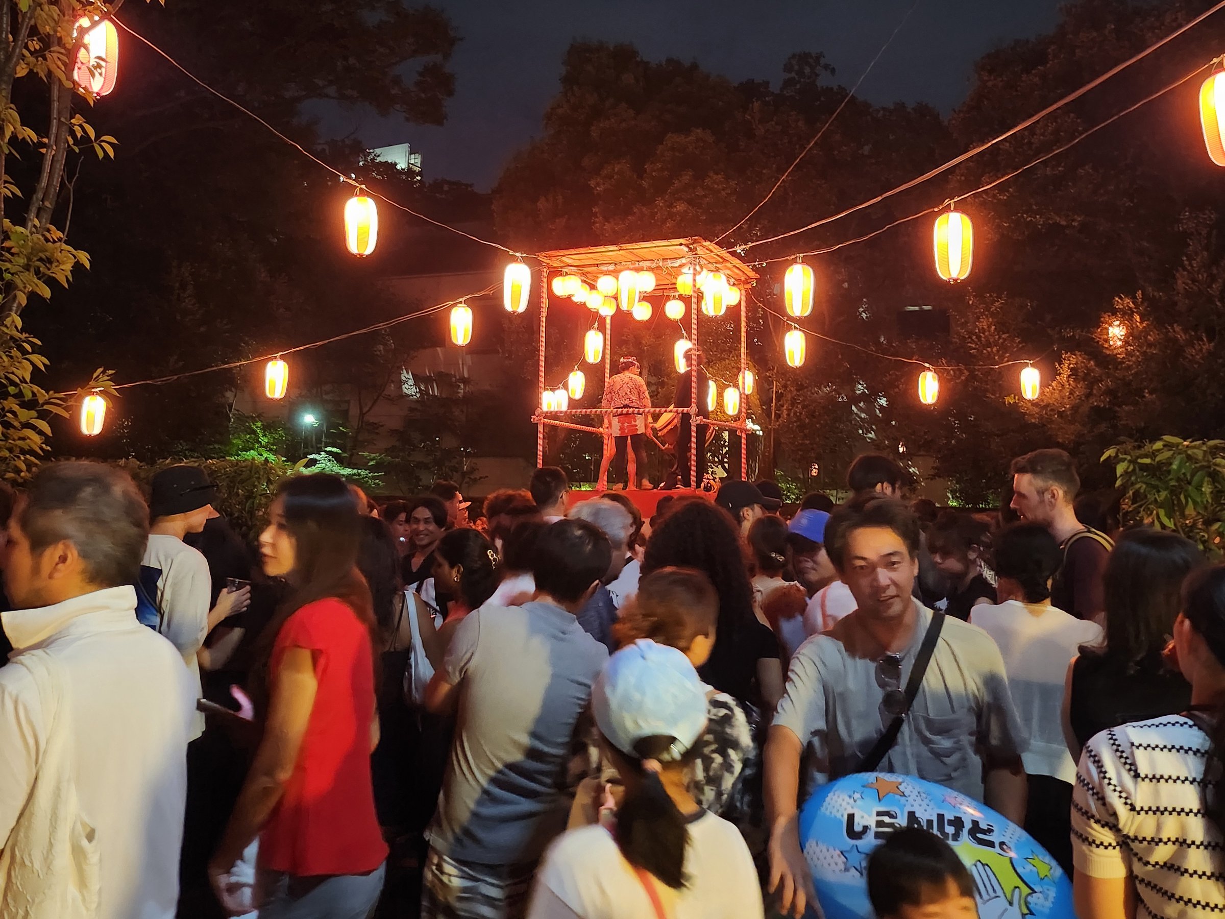A drummer on a yagura tower decorated with rows of glowing paper lanterns at a nighttime bon odori in Tokyo, with a crowd gathered around it.