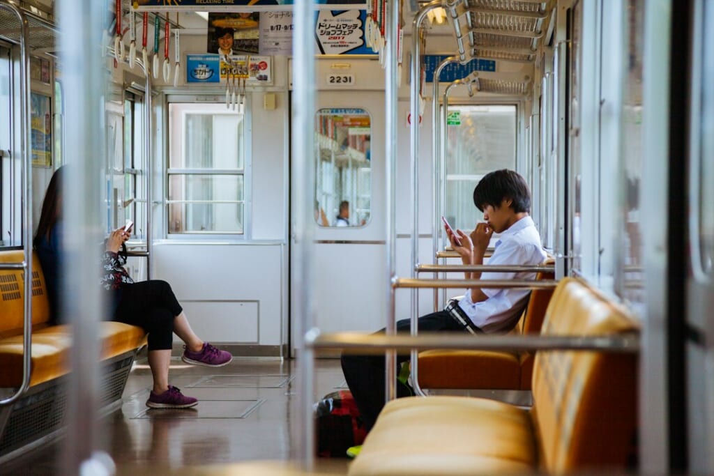Passengers seated inside a Japanese commuter train car with long bench seats and hanging straps.