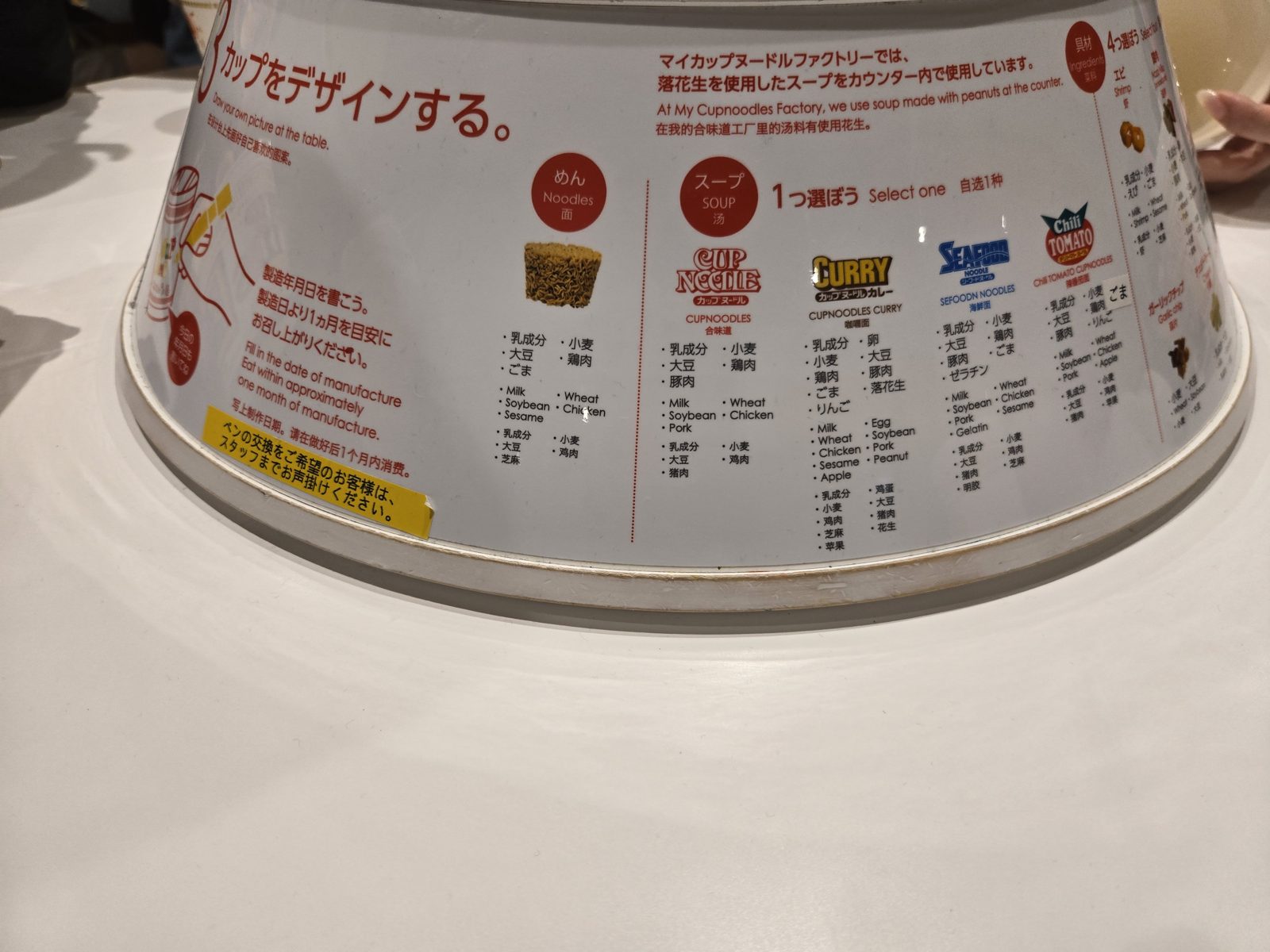 Sign on a drawing table at the My CUPNOODLES Factory showing the four soup choices, Cup Noodle, Curry, Seafood and Chili Tomato, with allergen information in Japanese and English.