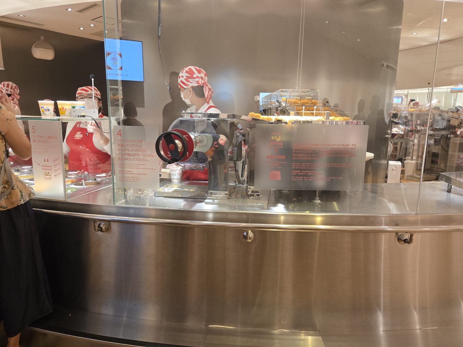 Staff in red-and-white bandanas working behind the stainless steel counter where toppings are added and cups are sealed at the My CUPNOODLES Factory.