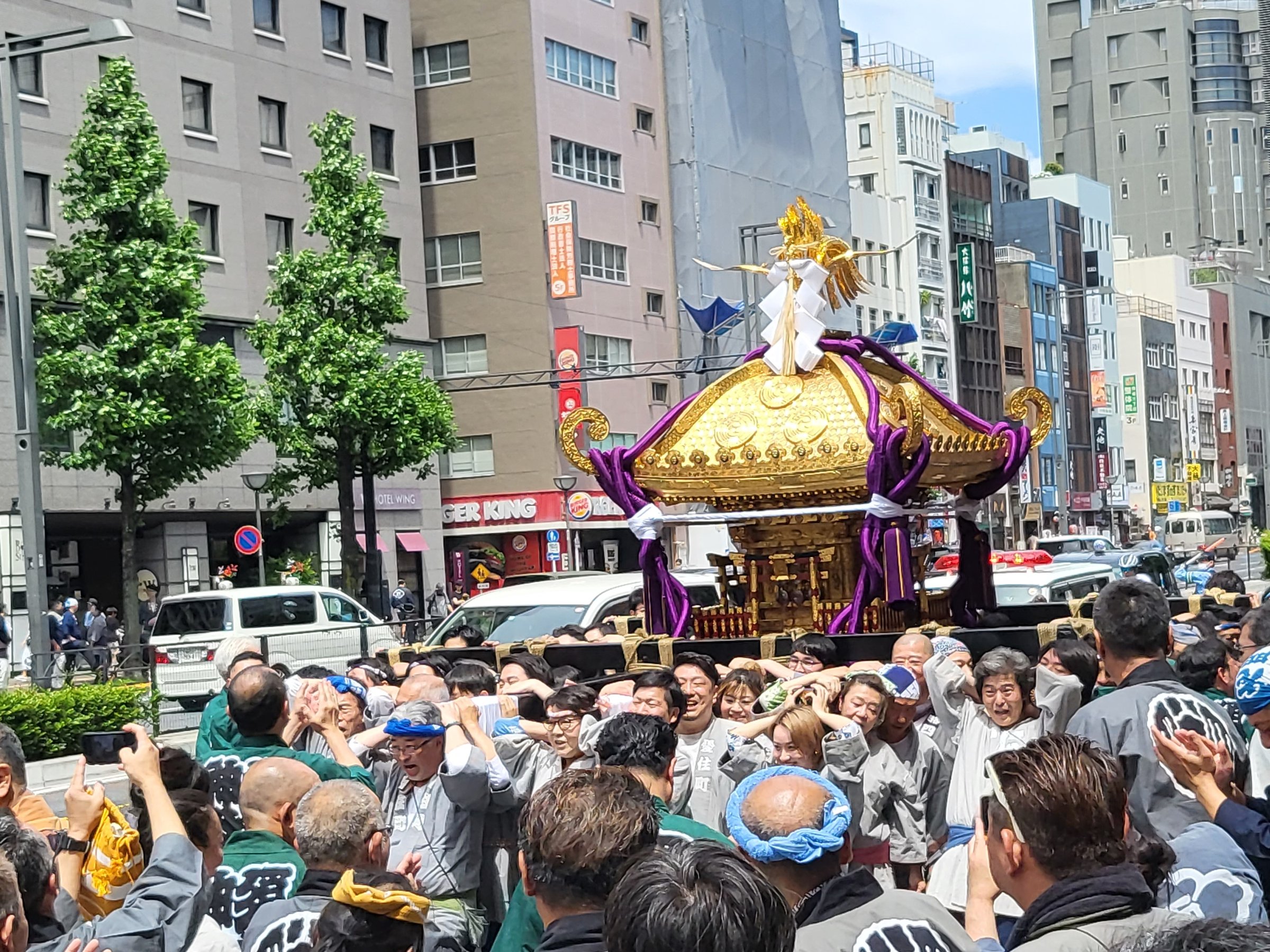 A large golden mikoshi portable shrine carried through a city street by bearers in matching happi coats, surrounded by onlookers taking photos.
