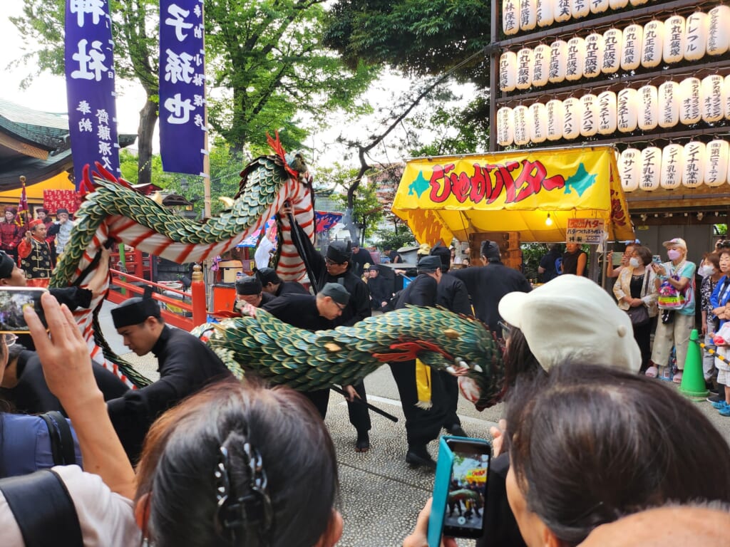 A dragon dance performed by men in black in a Tokyo shrine's grounds during a June festival, with festival stalls and rows of paper lanterns behind.