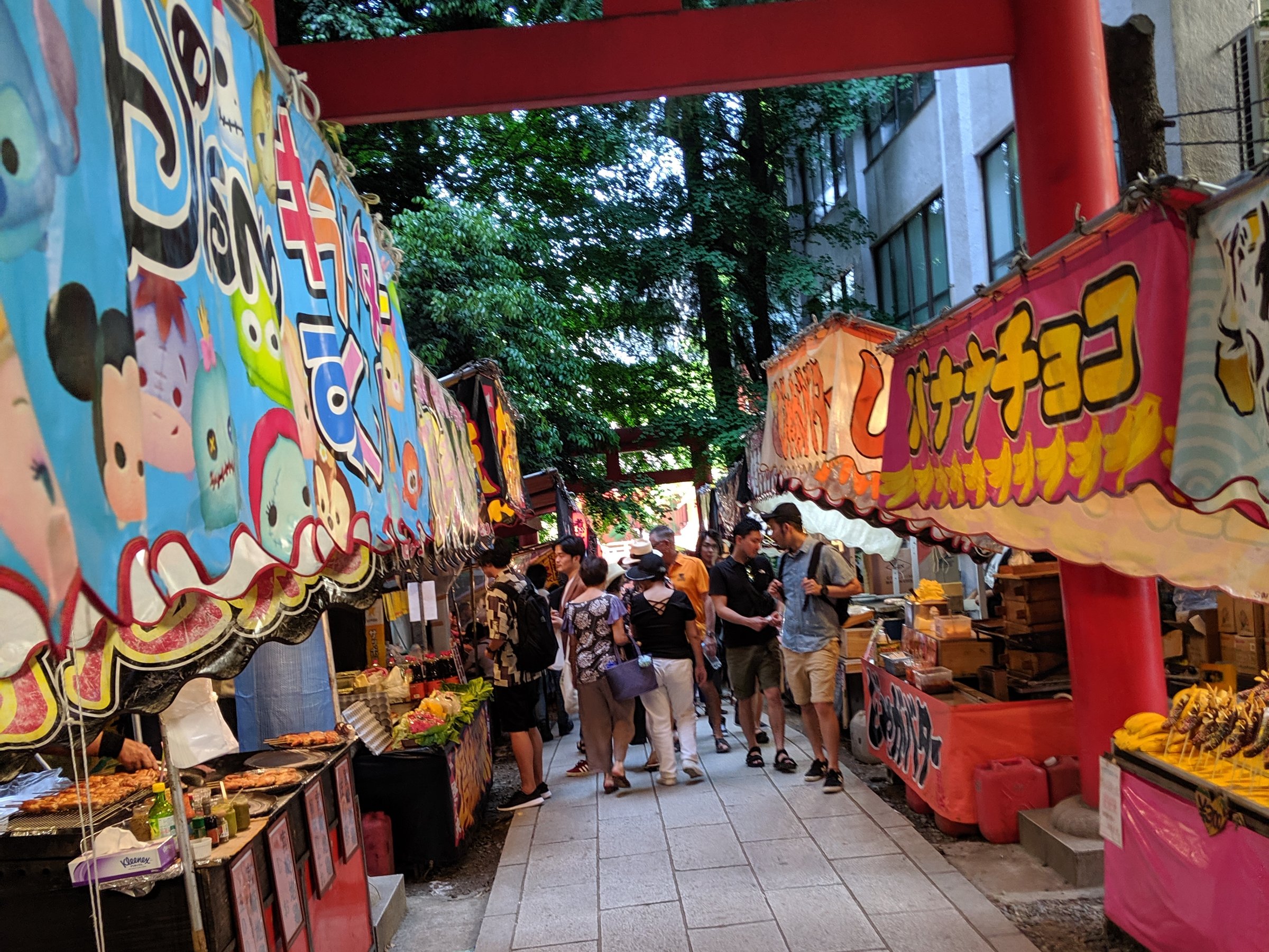 Festival stalls with colorful banners lining a stone path beneath a red torii gate at a Tokyo shrine.